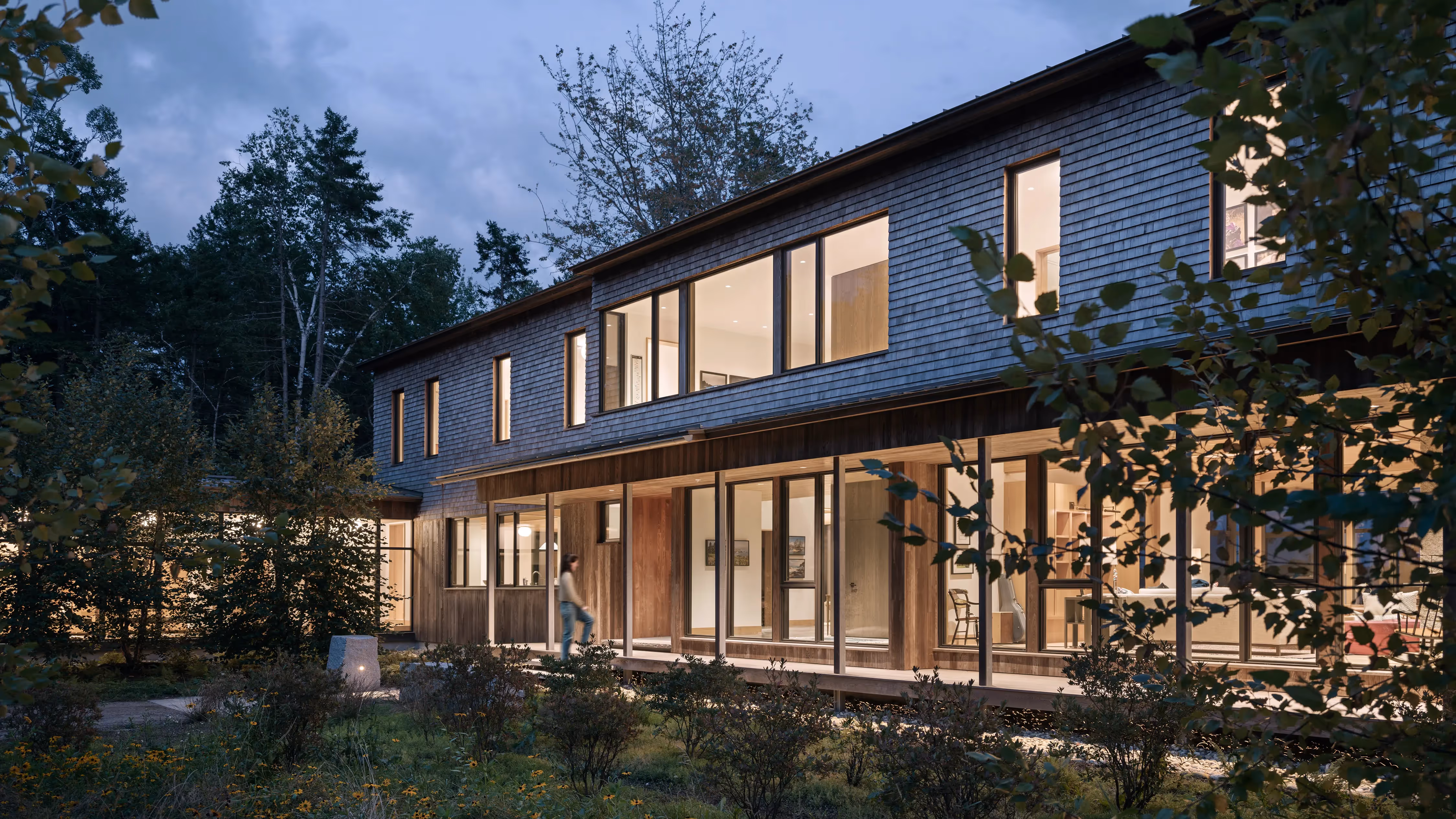 Modern two-story house with large windows nestled in wooded landscape at dusk