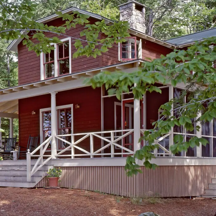 Red wooden cabin with white porch and railing, surrounded by green trees