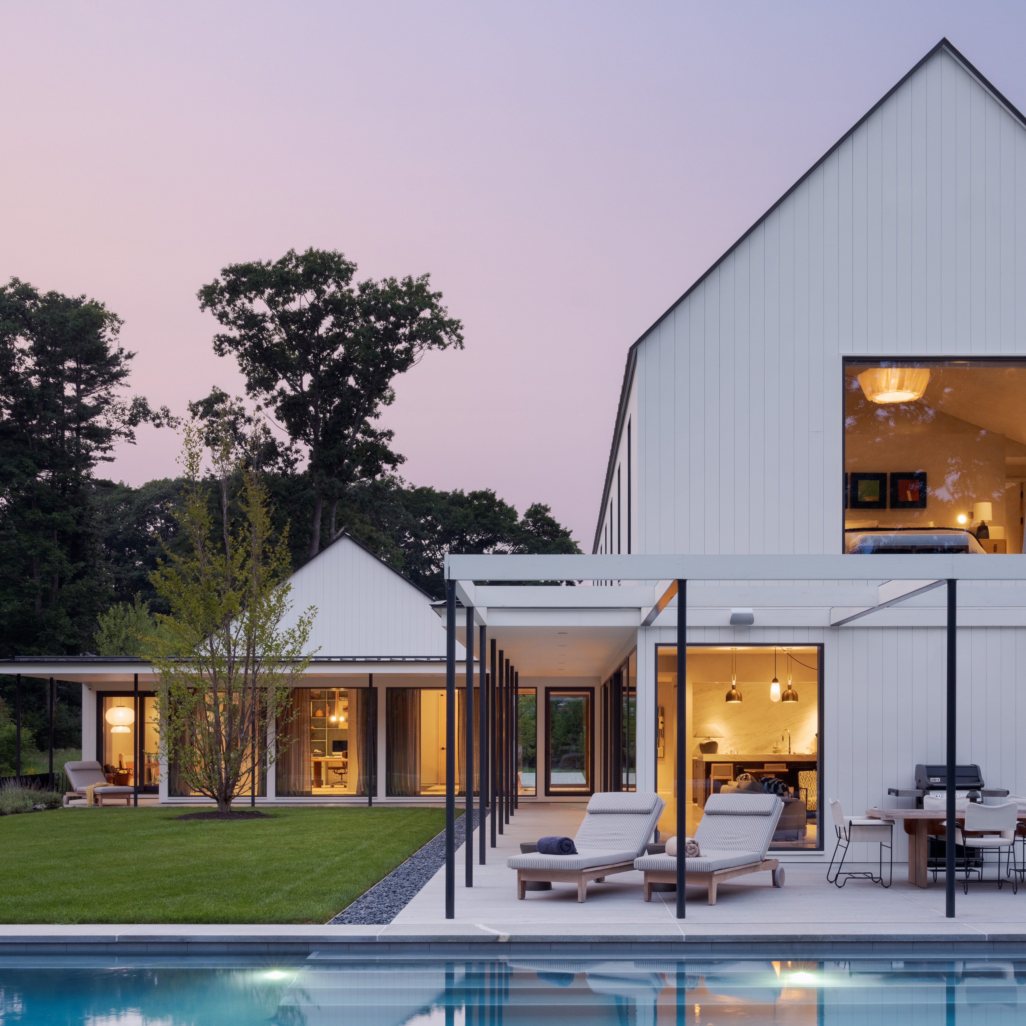 Modern white barn-style house with pool at twilight, surrounded by trees