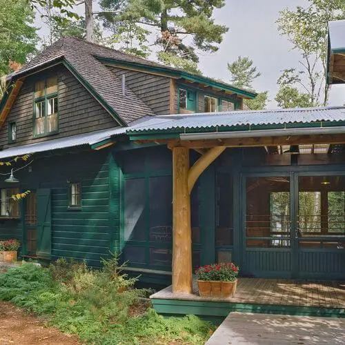 Rustic green wooden cabin with porch, surrounded by pine trees and greenery