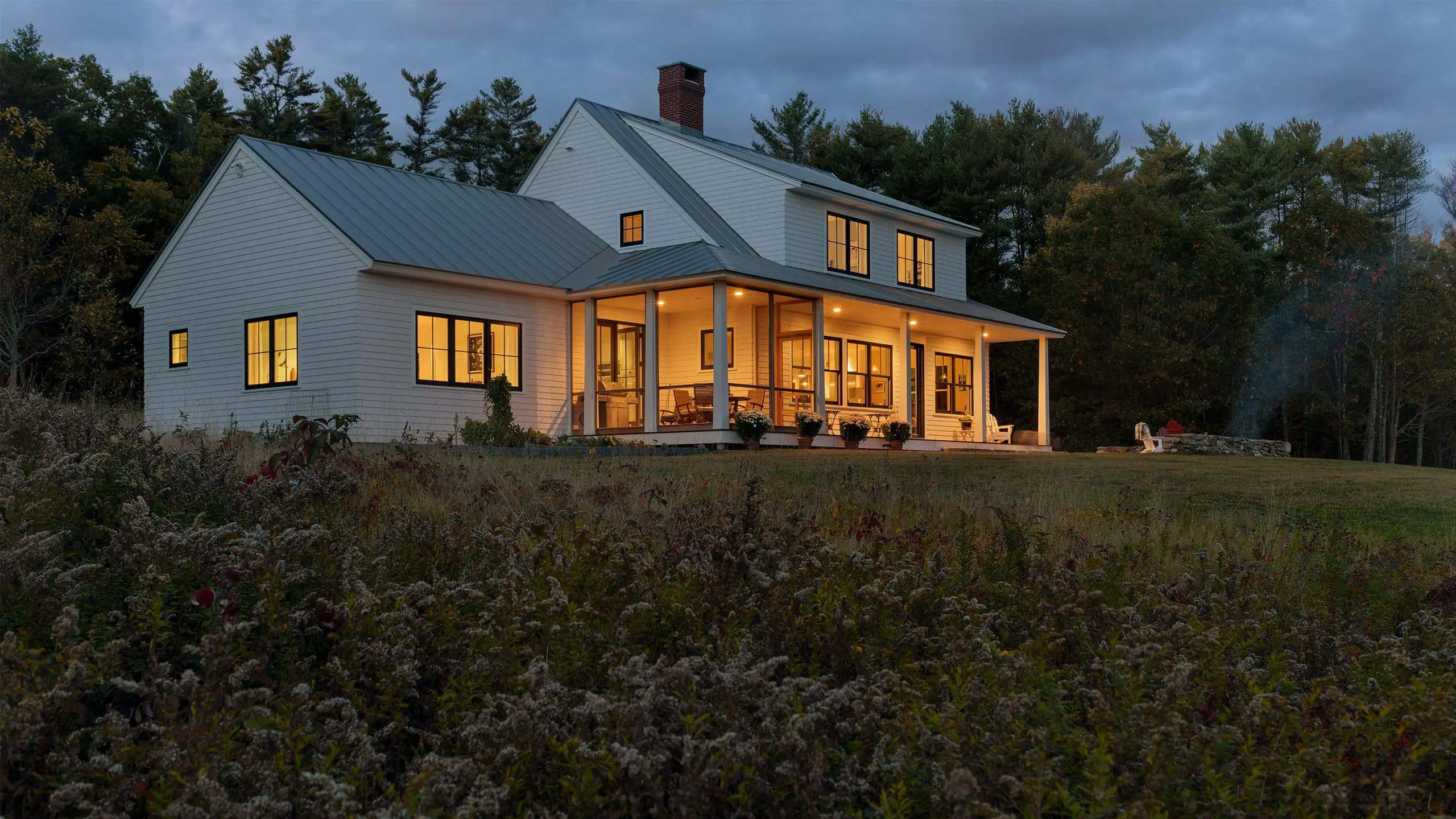 Warm, glowing white farmhouse with porch at dusk surrounded by pine trees