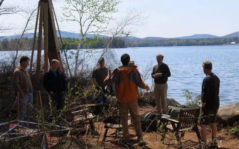 Group of people gathered by lake with mountains, discussing near rustic wooden structure