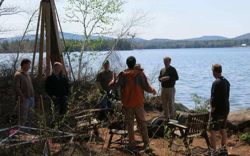 Group of people gathered by lake with mountains, discussing near rustic wooden structure