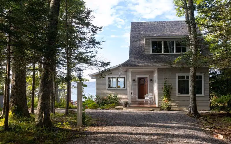 Coastal cottage nestled in pine forest with gravel driveway and white porch