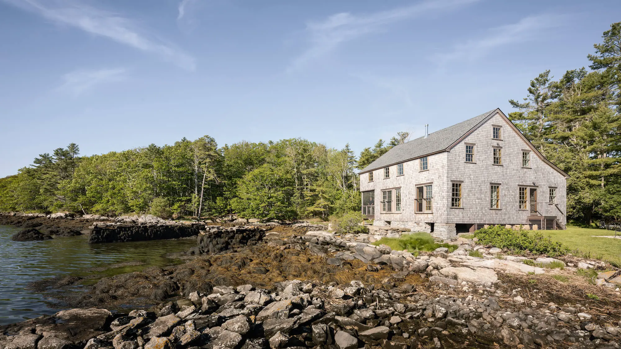 Shingle-clad house on rocky coastline surrounded by pine trees and water