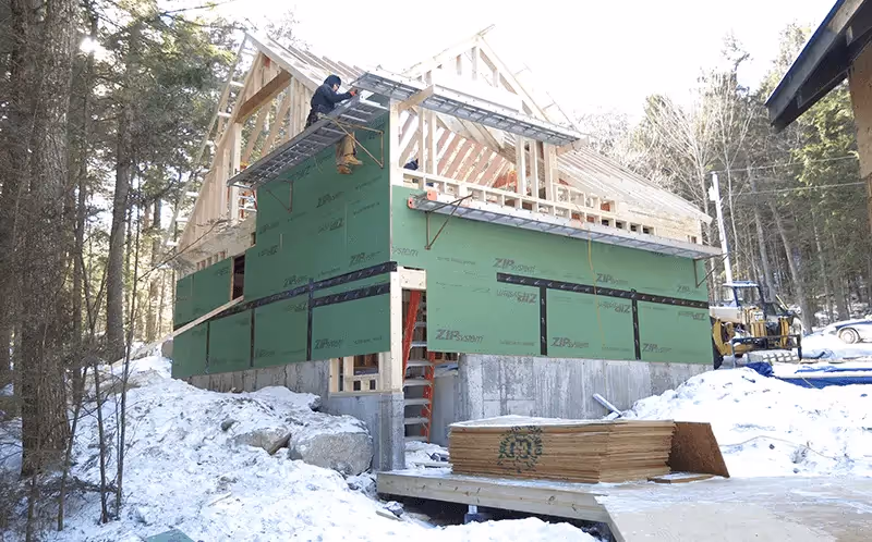 House under construction in snowy wooded area with green building wrap