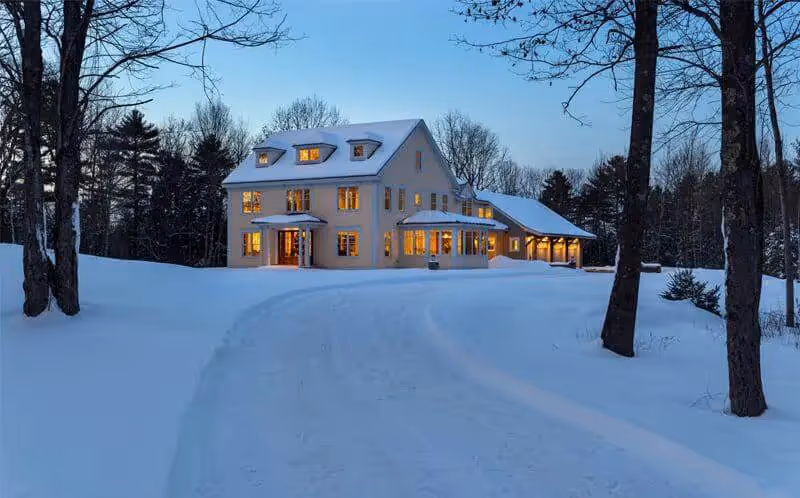 Warm-lit white house surrounded by snowy trees on a winter evening