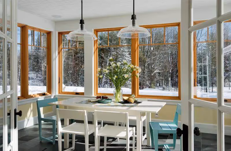 Bright sunroom with white table, teal chairs, and snowy forest view