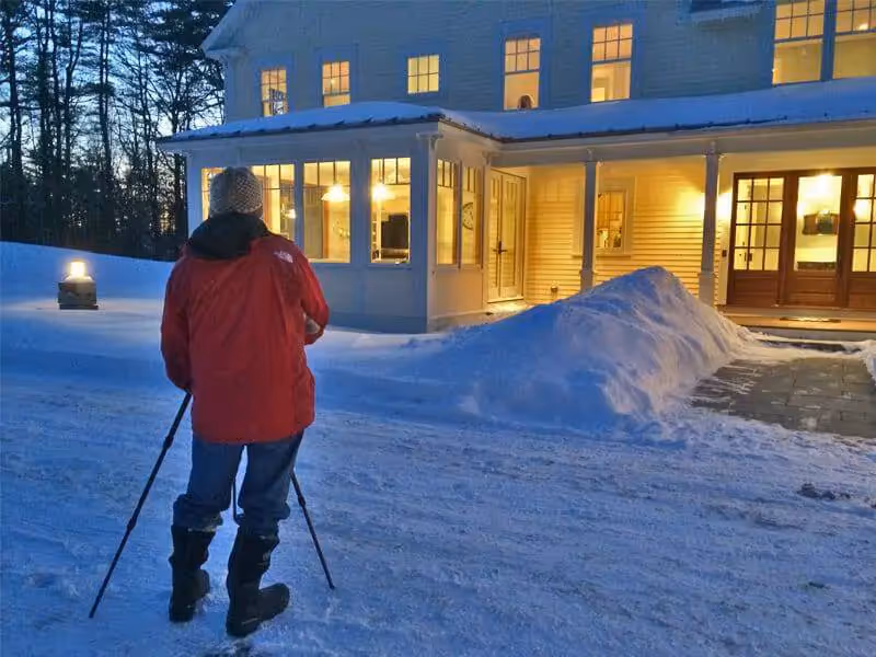 Person in red jacket shoveling snow near warm, lit house at dusk