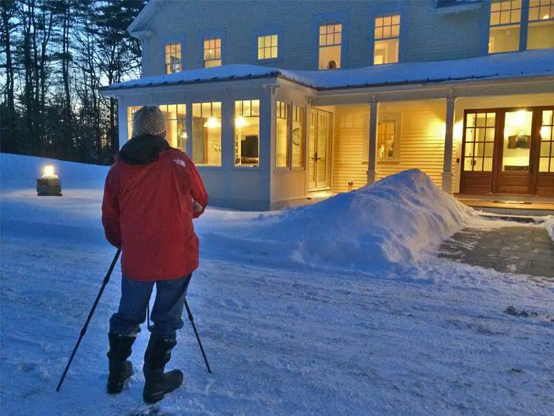 Person in red jacket shoveling snow near warm, lit house at dusk