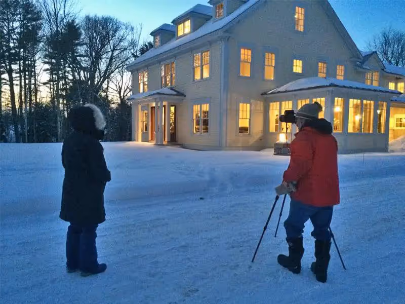 Photographer with tripod captures warm-lit house during snowy winter evening