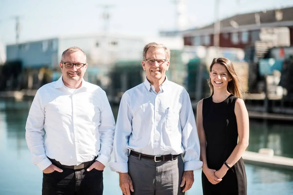 Three professionals smiling together by waterfront with industrial buildings