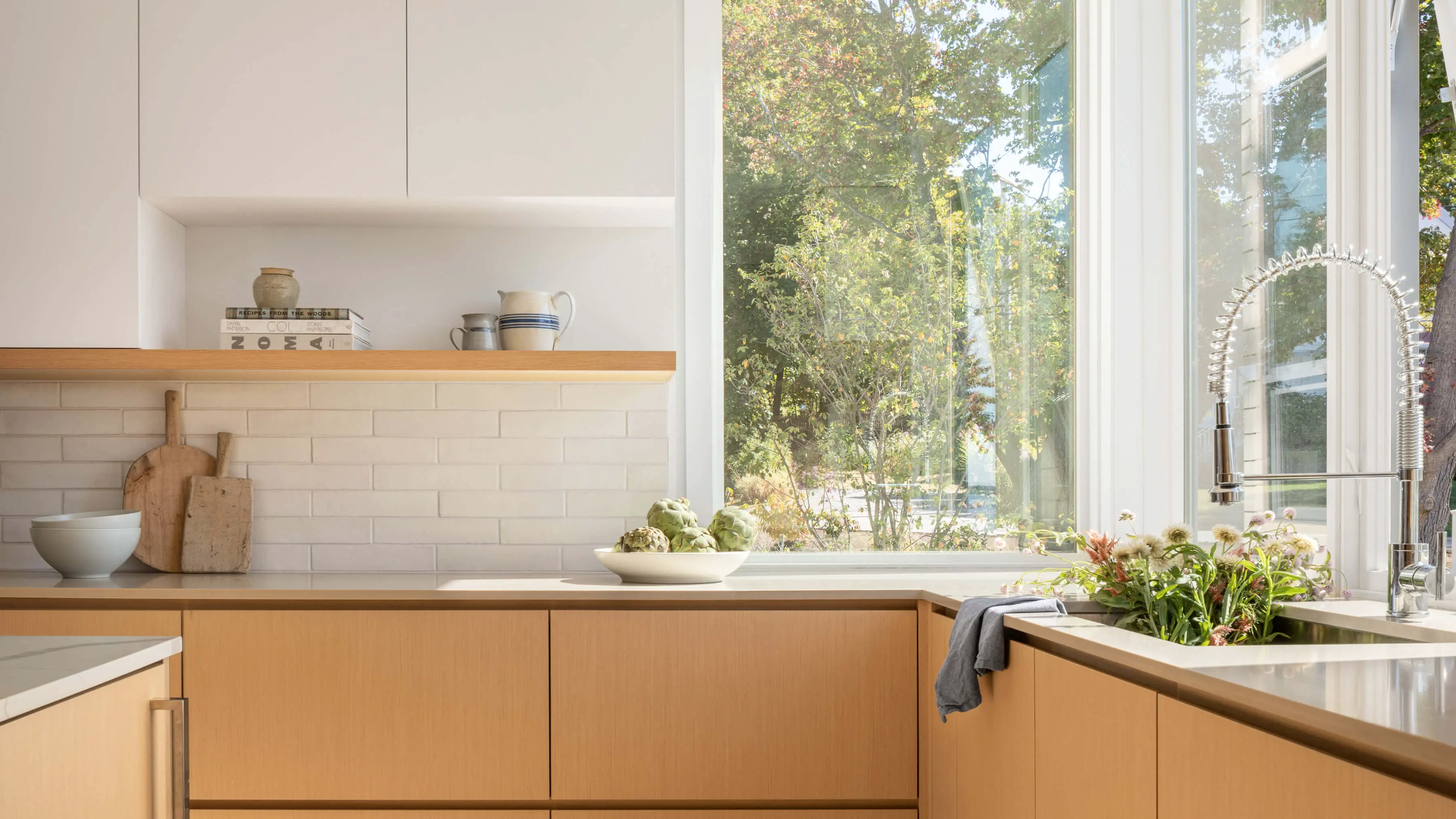 Modern kitchen with wooden cabinets, white tiles, and large windows overlooking trees
