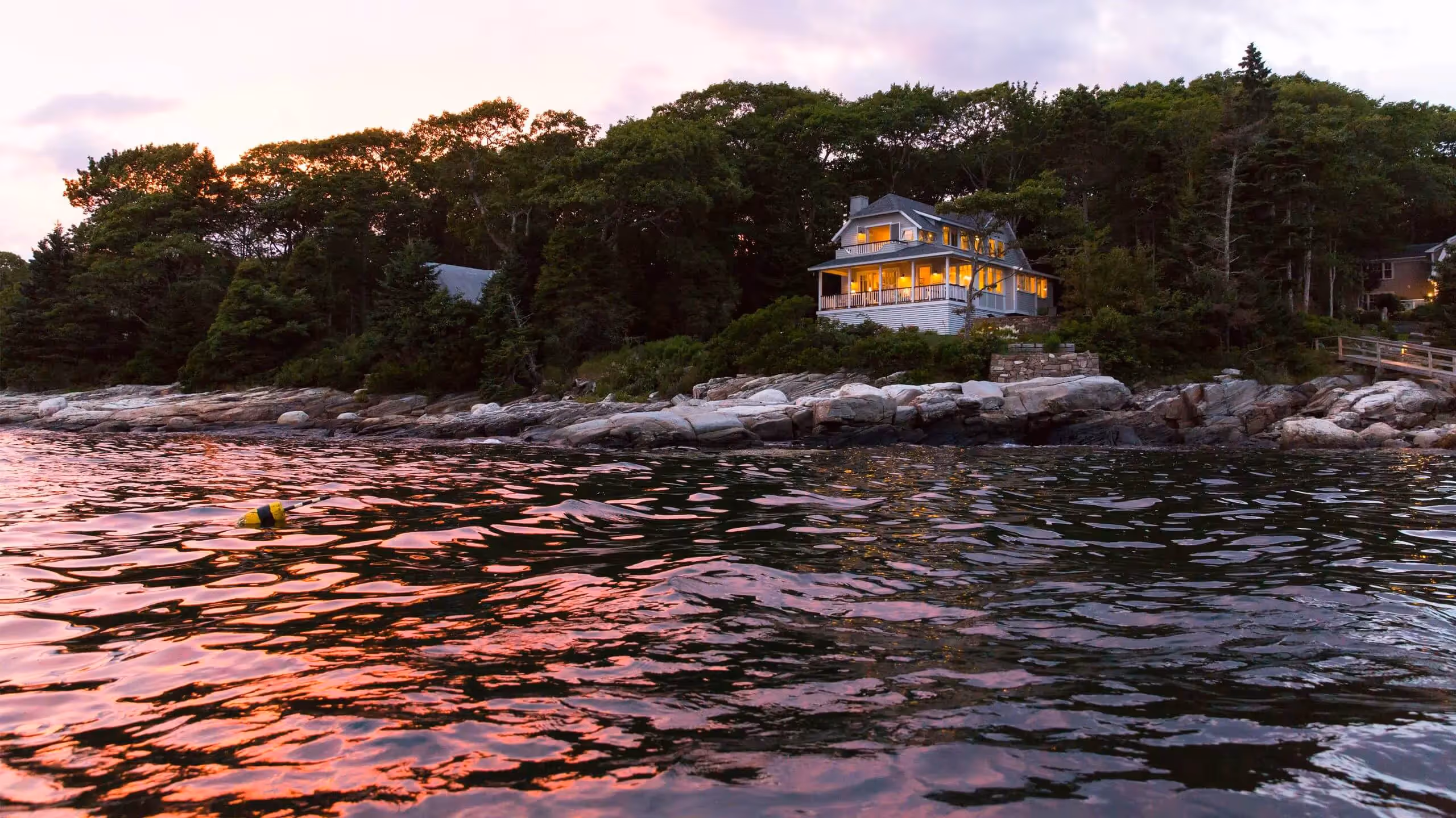 Glowing coastal cottage on rocky shore at sunset with pink water reflections