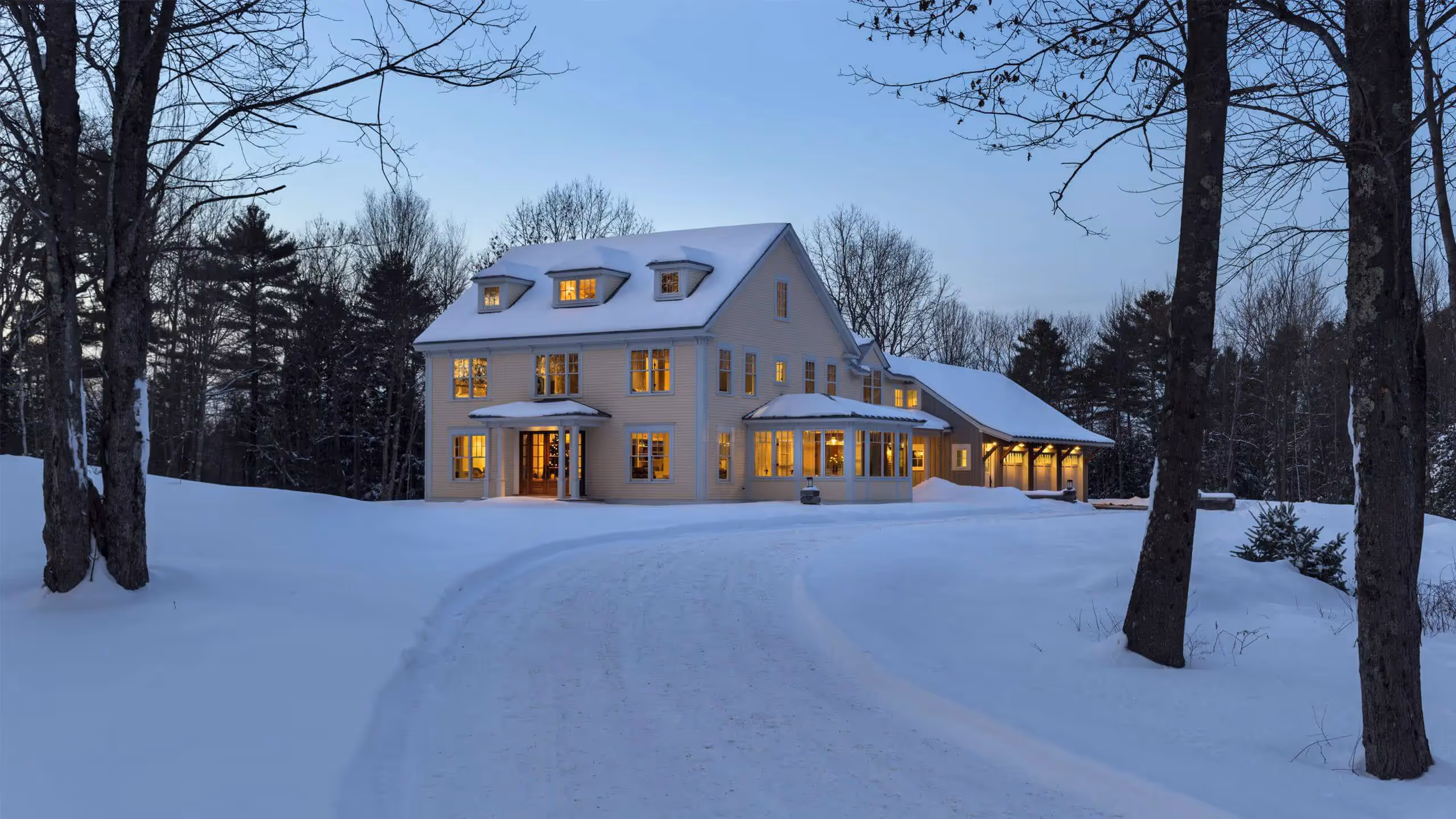Warm-lit two-story house surrounded by snowy trees at dusk