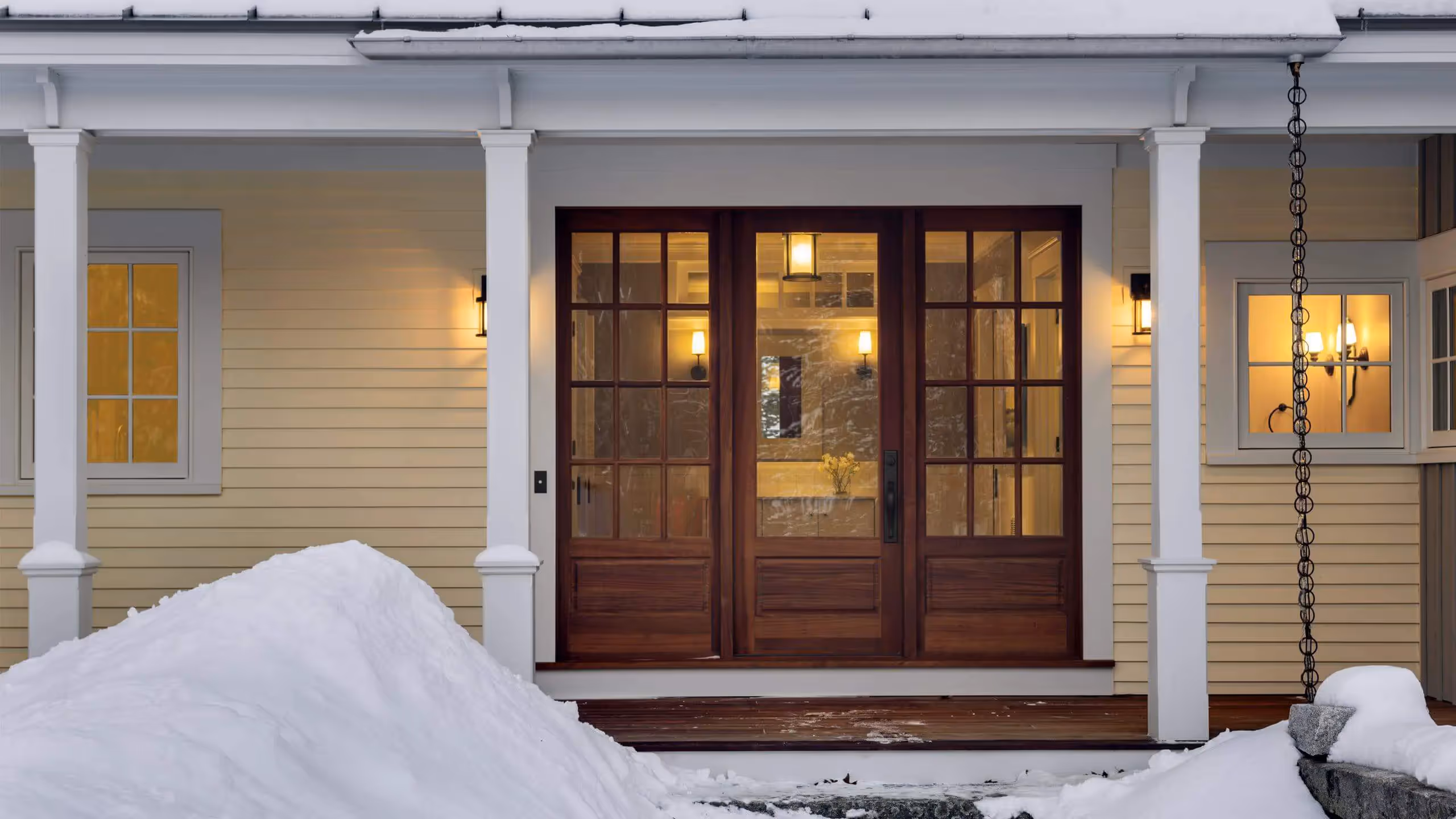 Wooden front door with glass panels, lit up at night, surrounded by snow