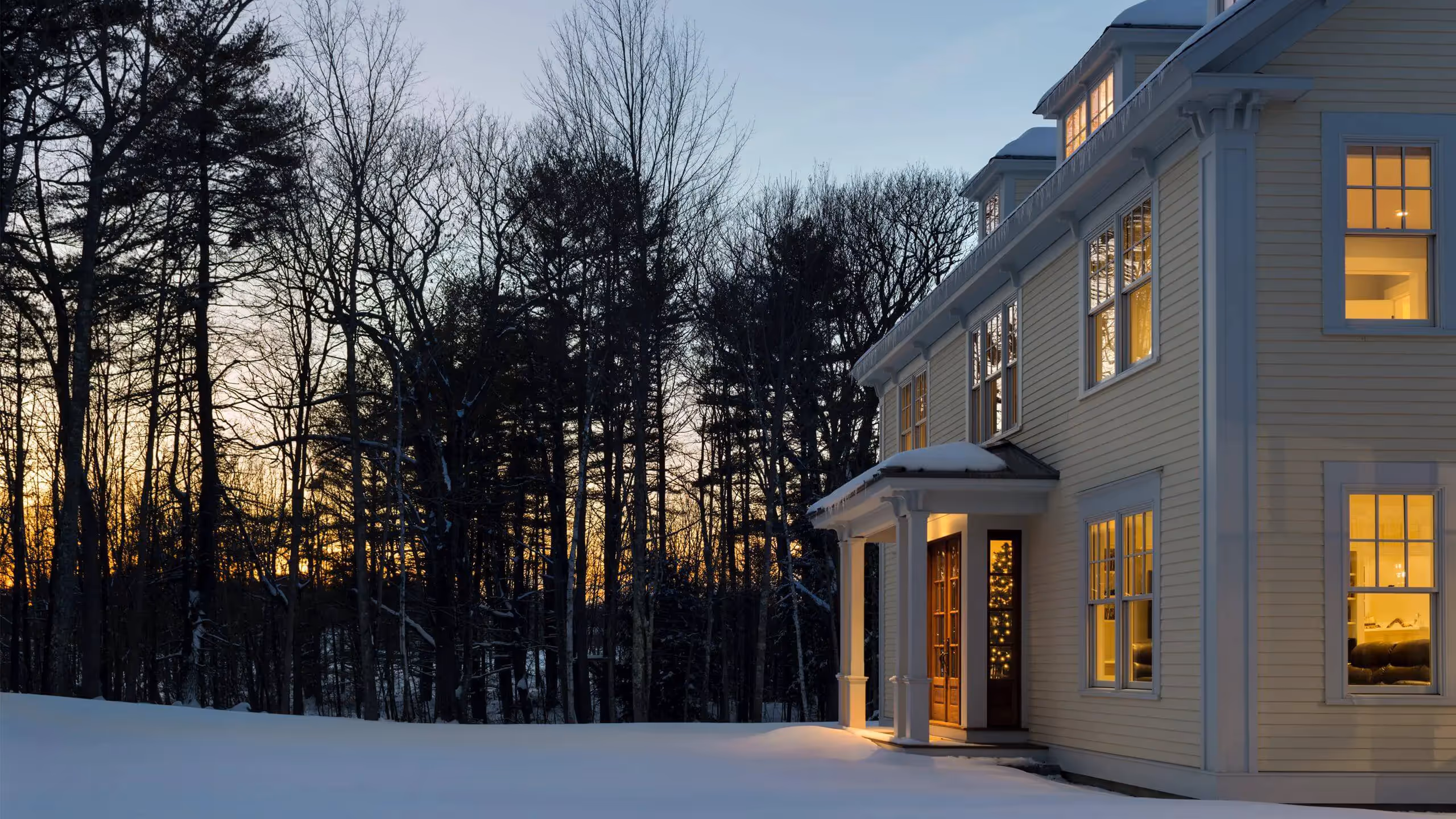 Warm glowing windows of white house at sunset in snowy winter forest
