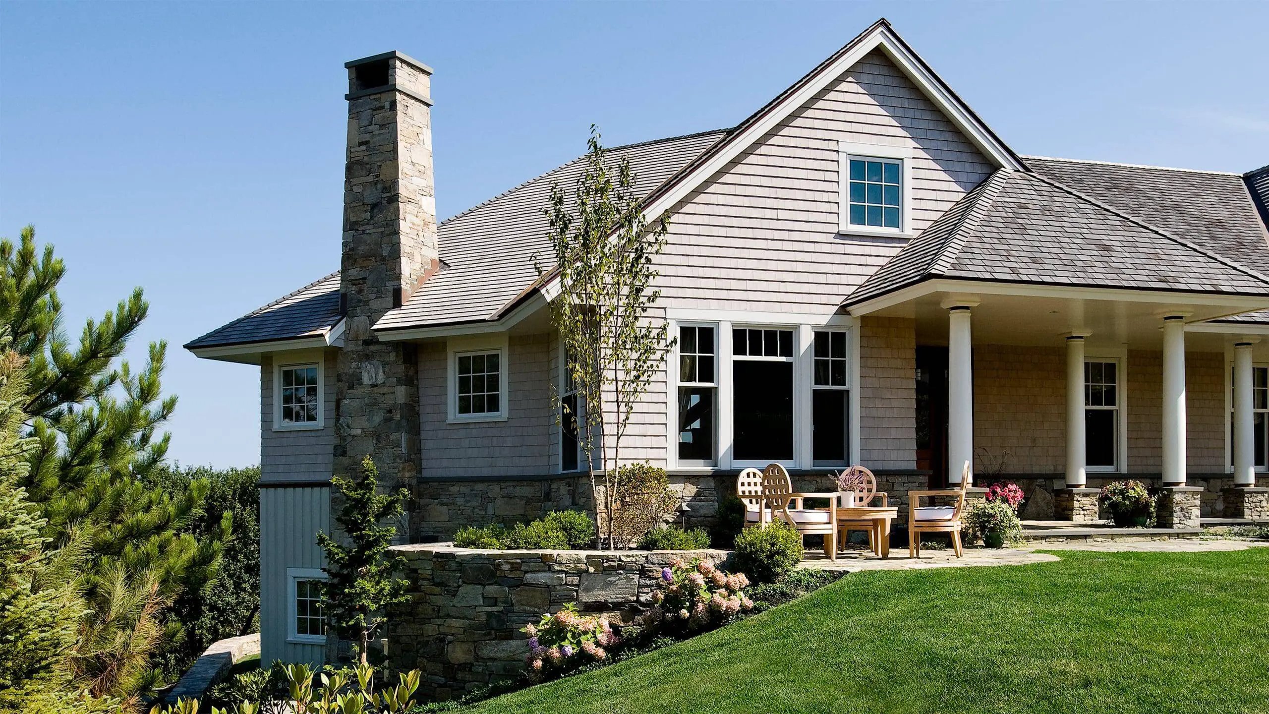 Stone and shingle coastal home with porch, landscaped yard, and wooden chairs