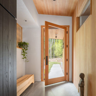 Open wooden door reveals forest path in minimalist entryway with wooden bench