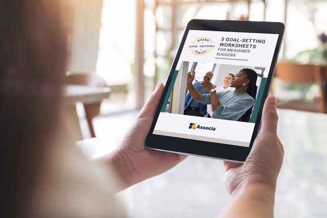 A person holding a tablet displaying an Associa eBook titled “5 Goal-Setting Worksheets for Measured Success” while sitting at a table in a bright indoor setting.