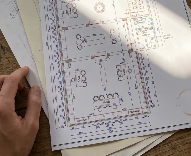 Hand holding an architectural floor plan showing tables, chairs, mirrors, mannequins, and an existing restroom.