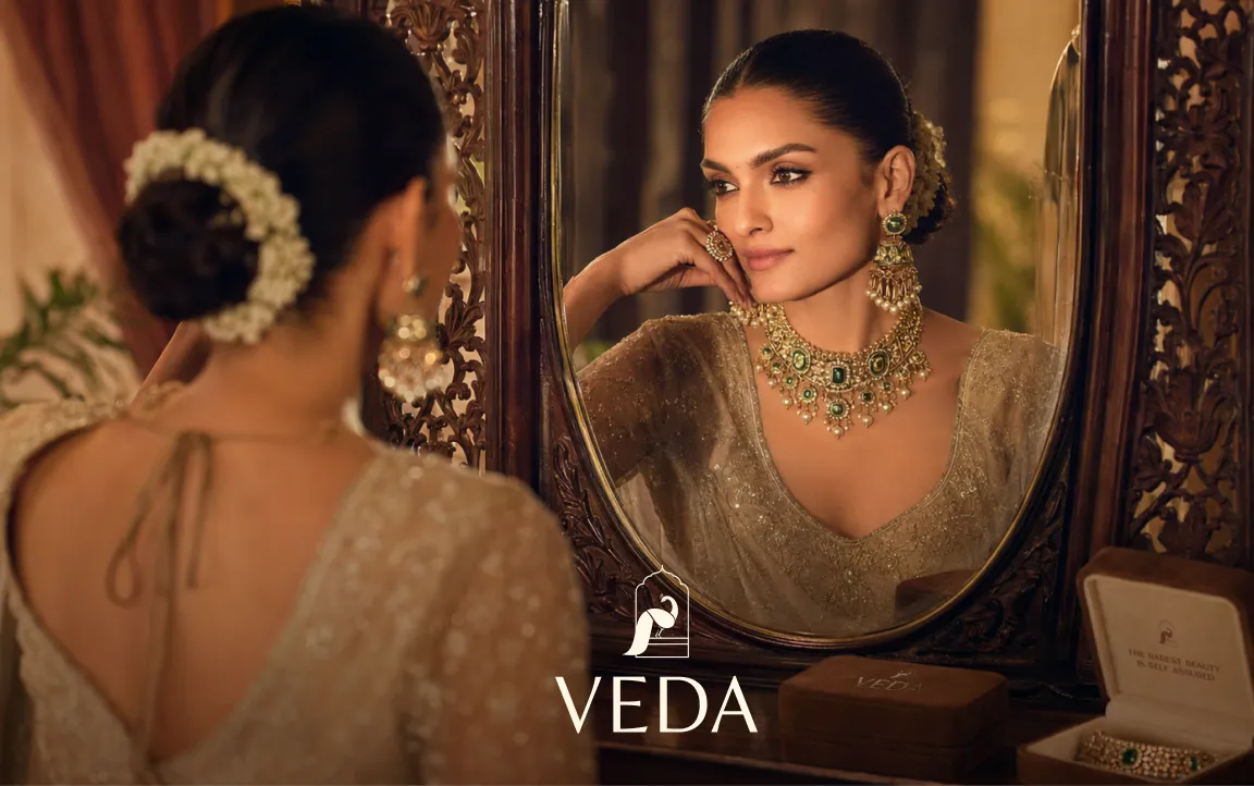 Woman with floral hair bun admiring her reflection wearing intricate green and gold jewelry at an ornate mirror.