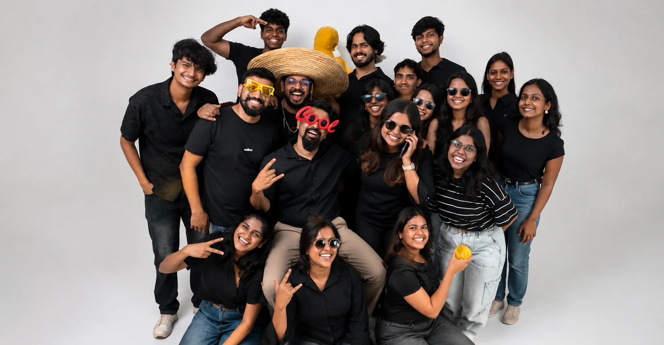Group of young people smiling and posing, some wearing sunglasses and fun glasses, in casual clothes.