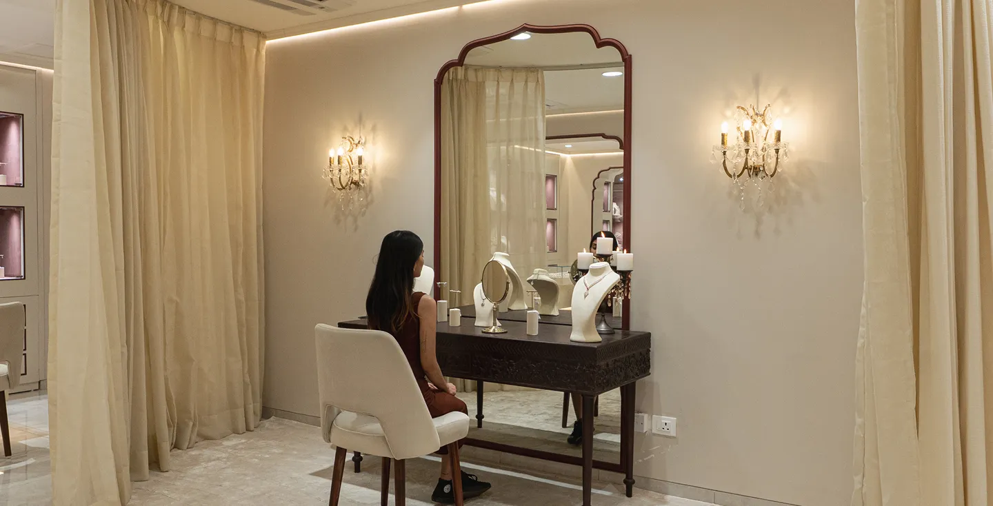Woman sitting on beige chair in front of wooden vanity with mirror and jewelry displays in an elegant room.