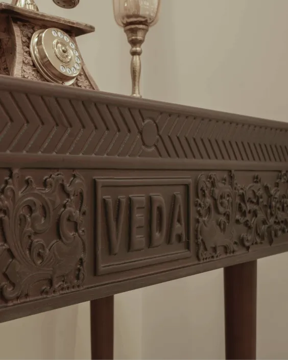 Brown wooden table with intricate carvings and the word VEDA on the side facing the camera.