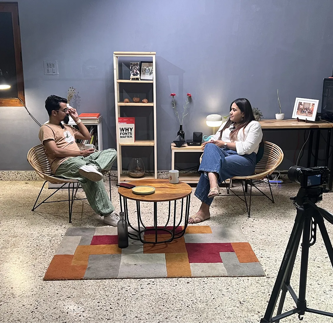 Two people sitting in rattan chairs facing each other with a round table and camera tripod in a living room.