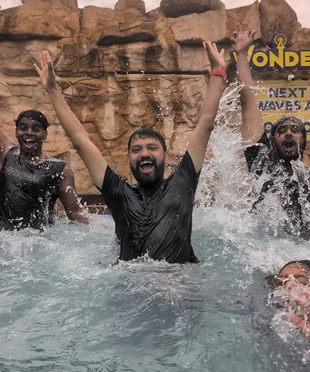 Four men in wet clothes cheering and splashing water in a pool with rocky background.