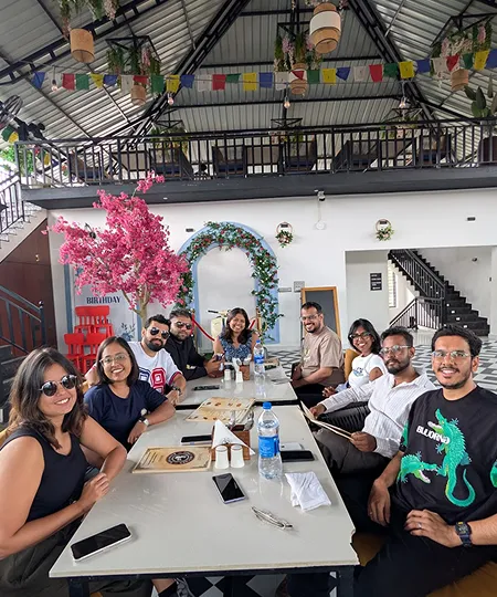 Group of eight friends smiling around a table in a bright indoor cafe with plants and colorful decor.