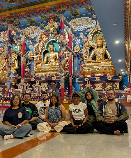Group of seven people sitting cross-legged in front of ornate golden Buddha statues in a colorful temple.