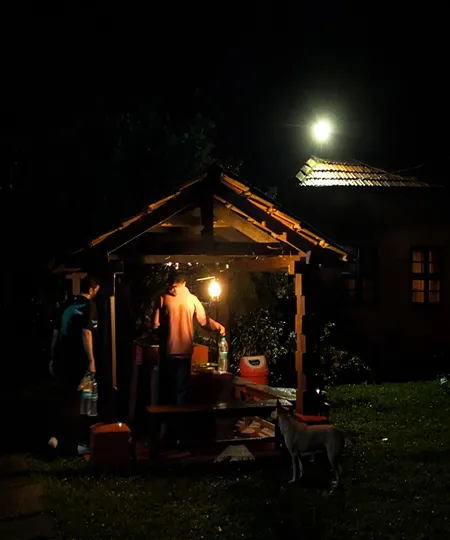 Two people at a wooden picnic shelter lit by a lantern and a distant bright light at night, with a dog nearby.