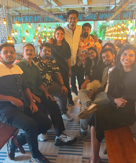 Group of nine young adults smiling and posing together in a warmly lit, decorated indoor space.