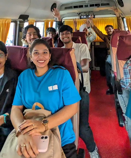 Group of smiling young people sitting and waving inside a bus with maroon seats and yellow curtains.