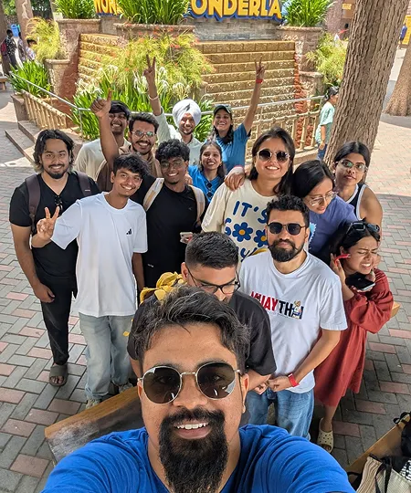 Group selfie of smiling young adults posing outdoors on paved pathway near a garden and tree.