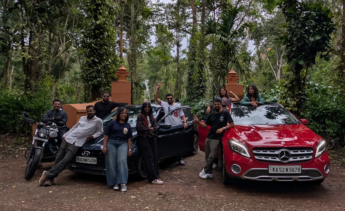 Group of people posing with a black car, red Mercedes SUV, and a motorcycle on a forest path.