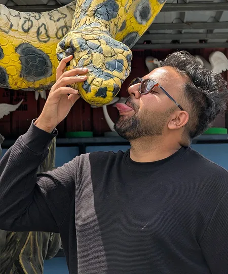 Man wearing sunglasses playfully licking the nose of a large yellow and gray snake statue.
