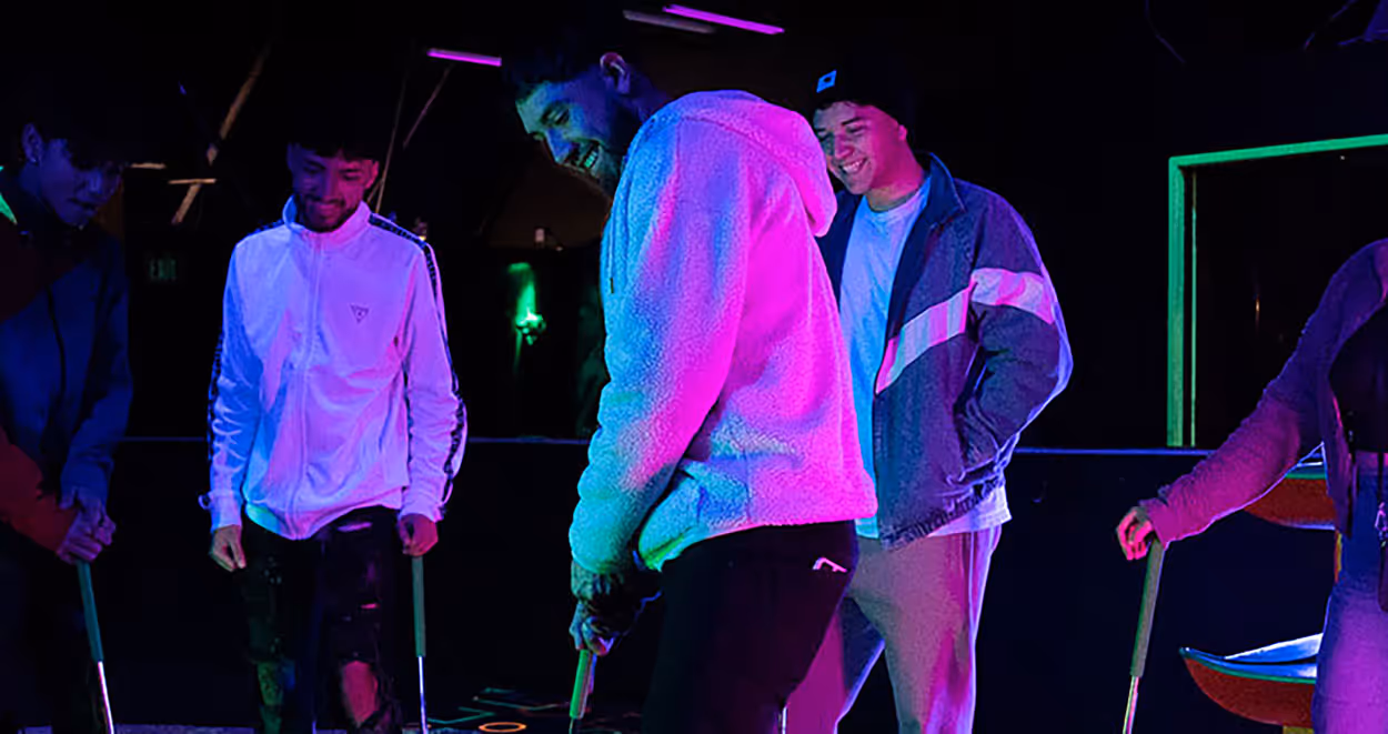 A group of young adults standing under neon lights, engaged in a social activity. They are dressed casually, with two individuals wearing hoodies. The setting appears to be indoors, with vibrant lighting creating a lively atmosphere.