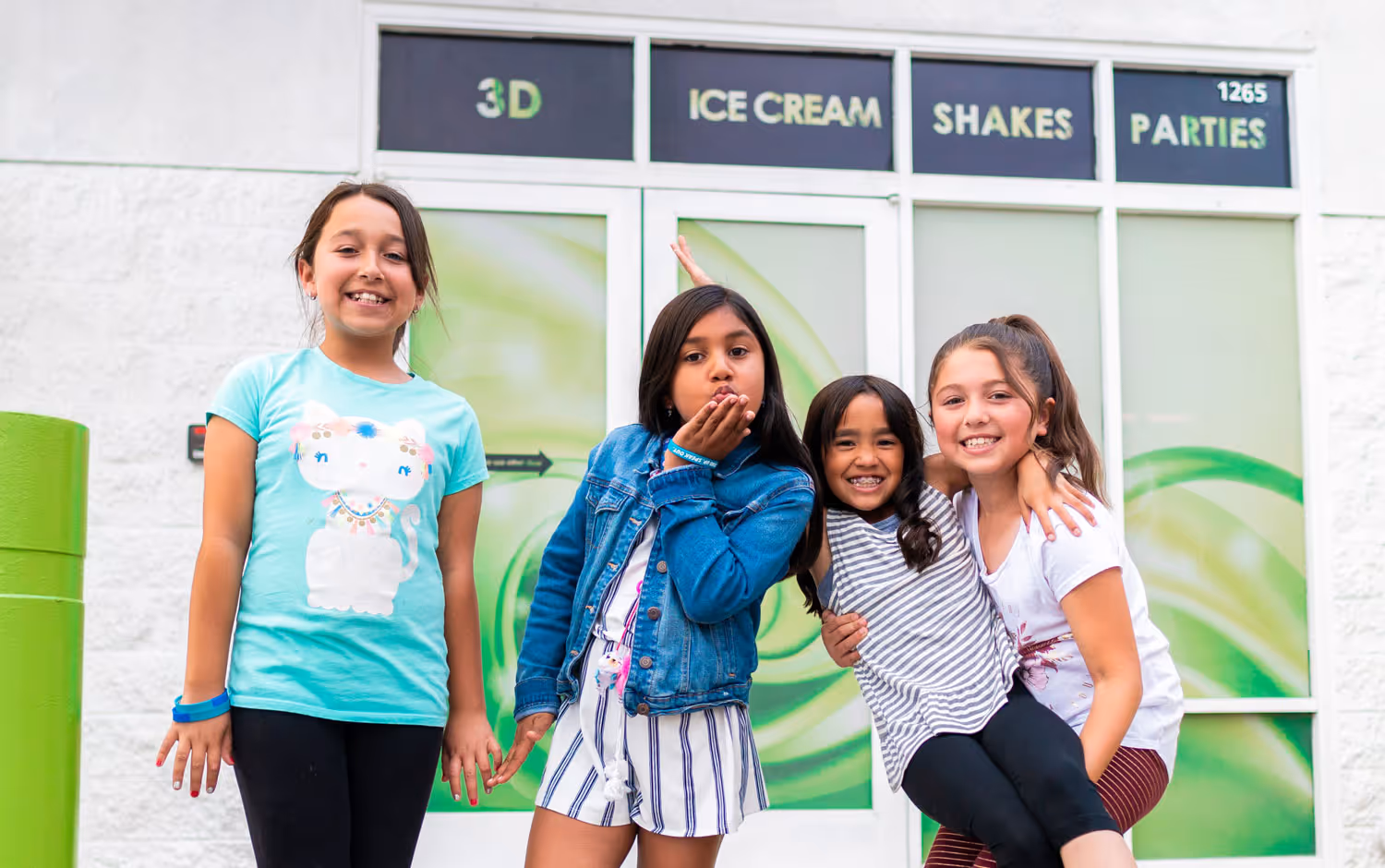 Four girls posing in front of a shop with a green and white design. The store's sign indicates it offers ice cream, shakes, and party services. The girls are smiling and wearing colorful clothing.