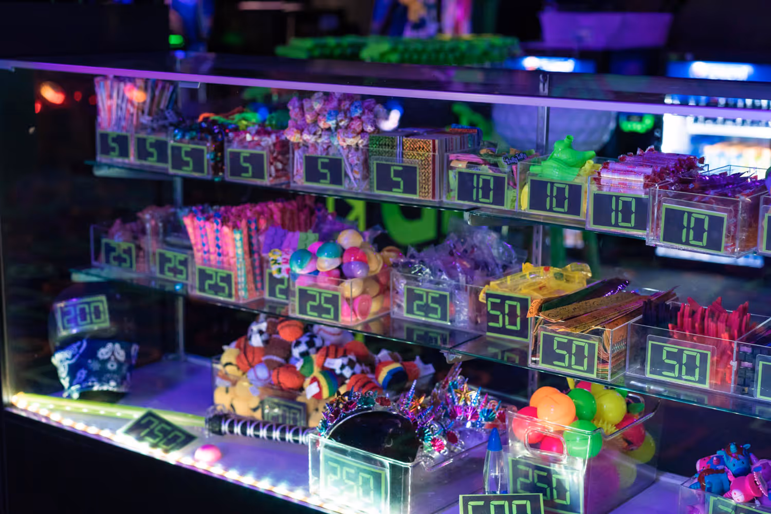 A brightly lit display case filled with colorful prize items and their point values. The items include small toys, candy, and other trinkets, each labeled with a price ranging from 5 to 200 points.