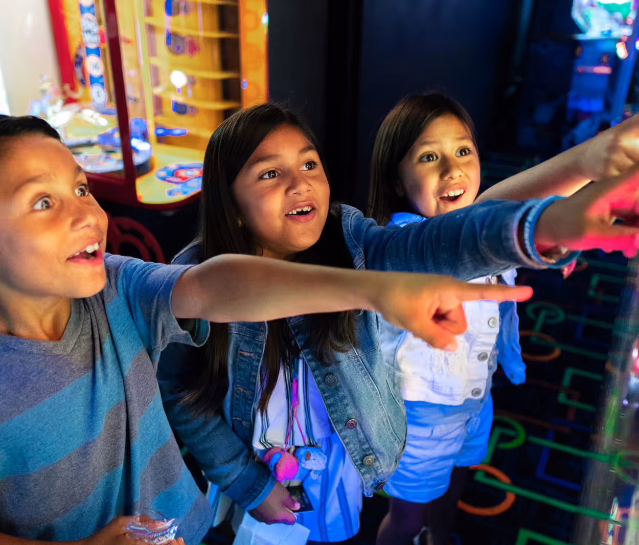 Three children excitedly pointing and looking at something in an arcade. The boys wear a striped shirt, while a girl wears a denim jacket. Colorful lights and arcade games are visible in the background.