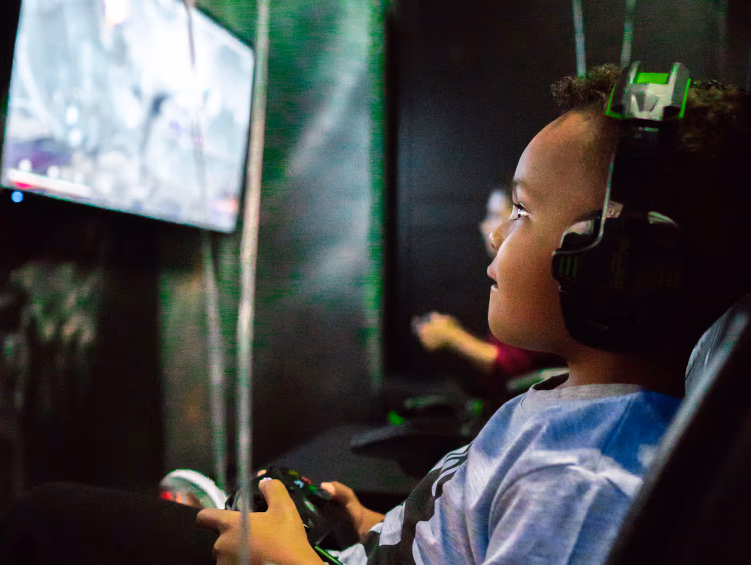 A child wearing a headset plays video games on a console, focused on the screen in a dimly lit gaming area. Another person is visible in the background, also gaming.