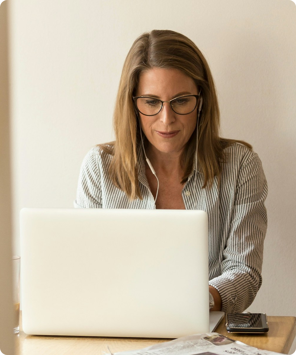 Woman wearing glasses and earphones, working on a laptop at a wooden table with a smartphone and newspaper.