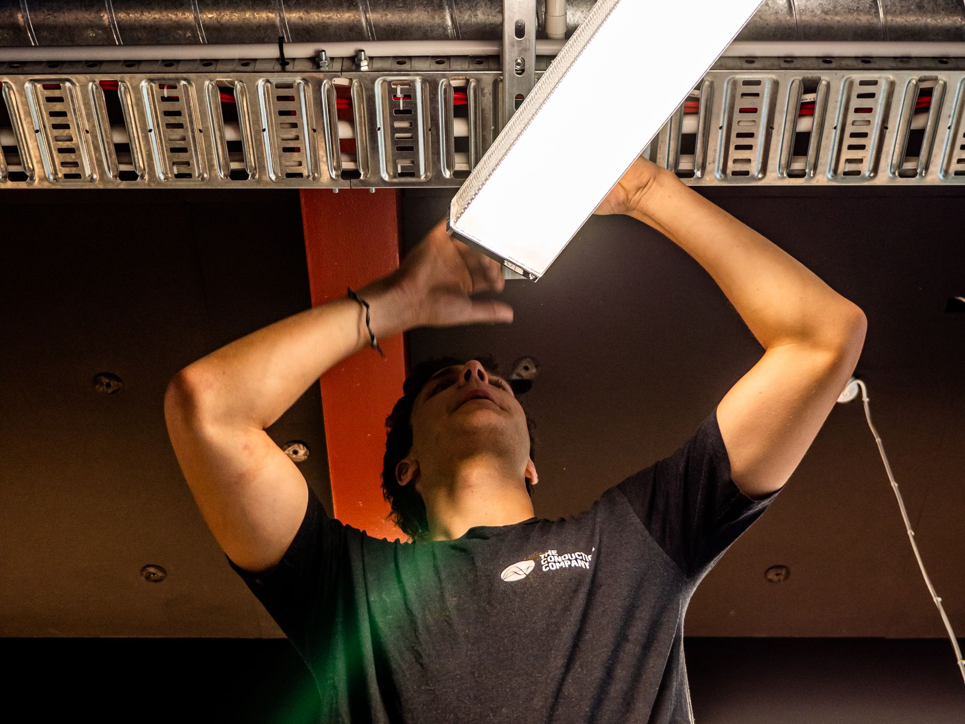 An electrician installing a linear LED light fitting on a commercial ceiling beside cable tray
