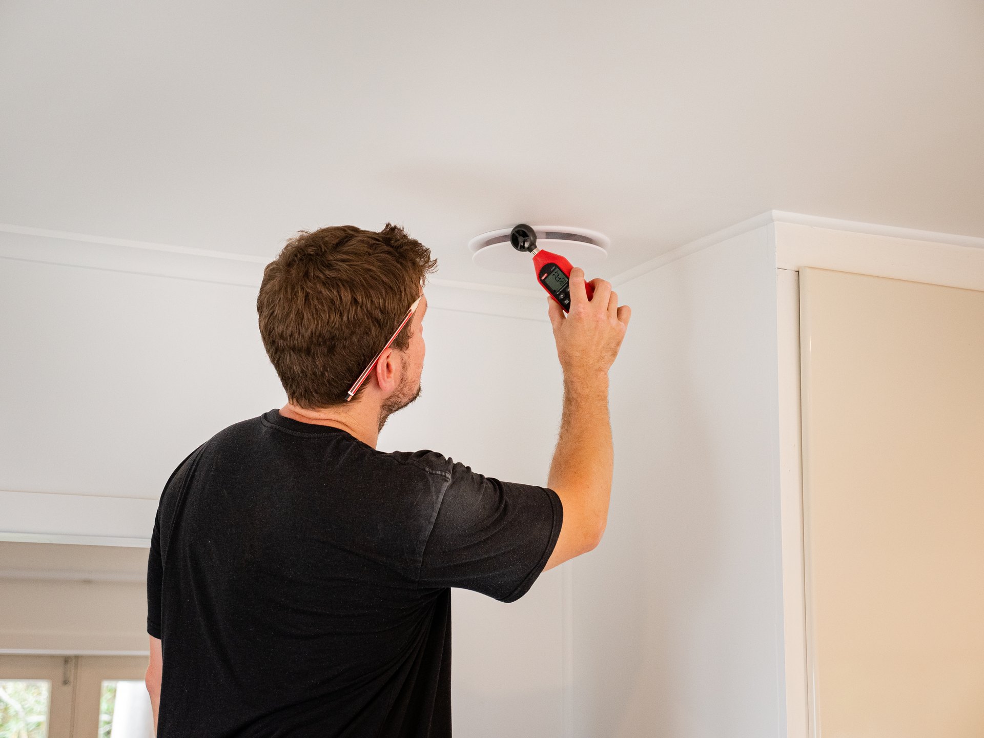 An electrician reaching up to work on a ceiling fitting in a plain, lived in room