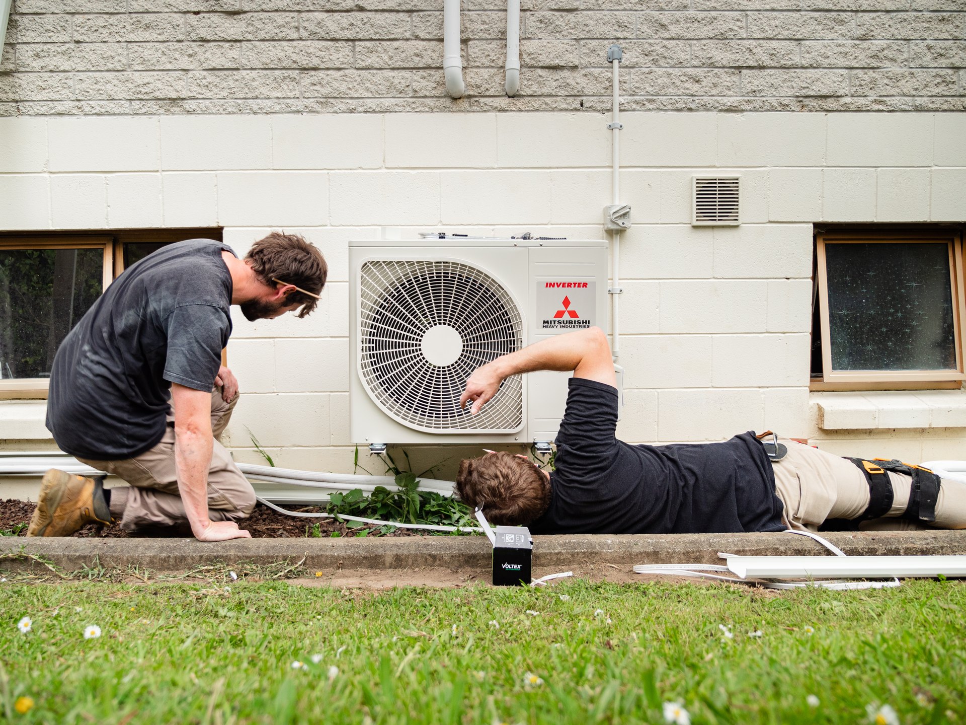 Two electricians working on a heat pump unit mounted on the outside wall of a house