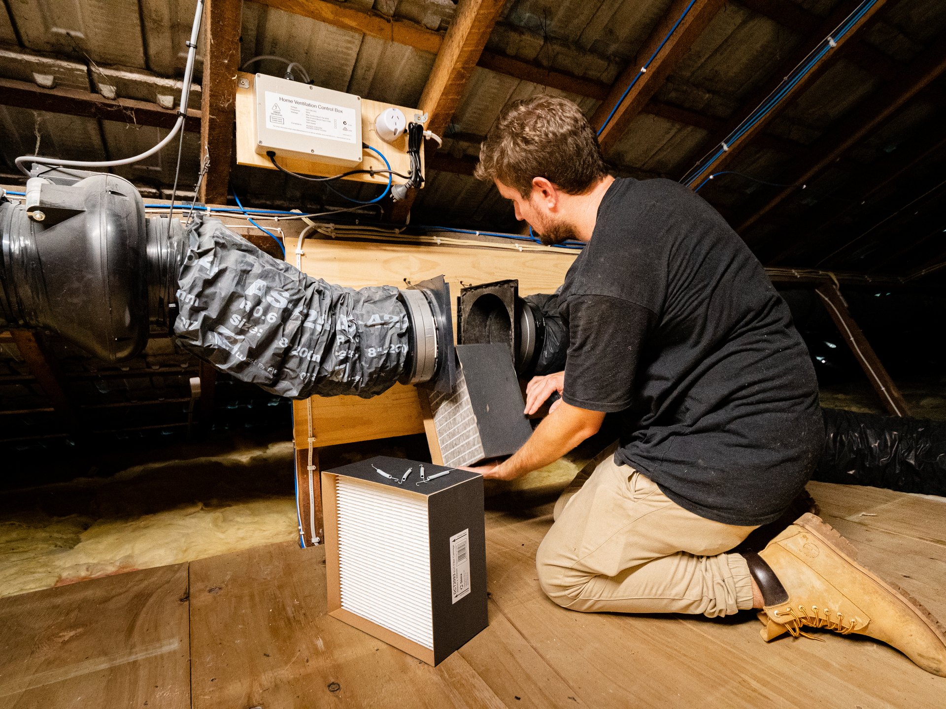 An electrician kneeling in a roof space, fitting a ventilation unit to ducting