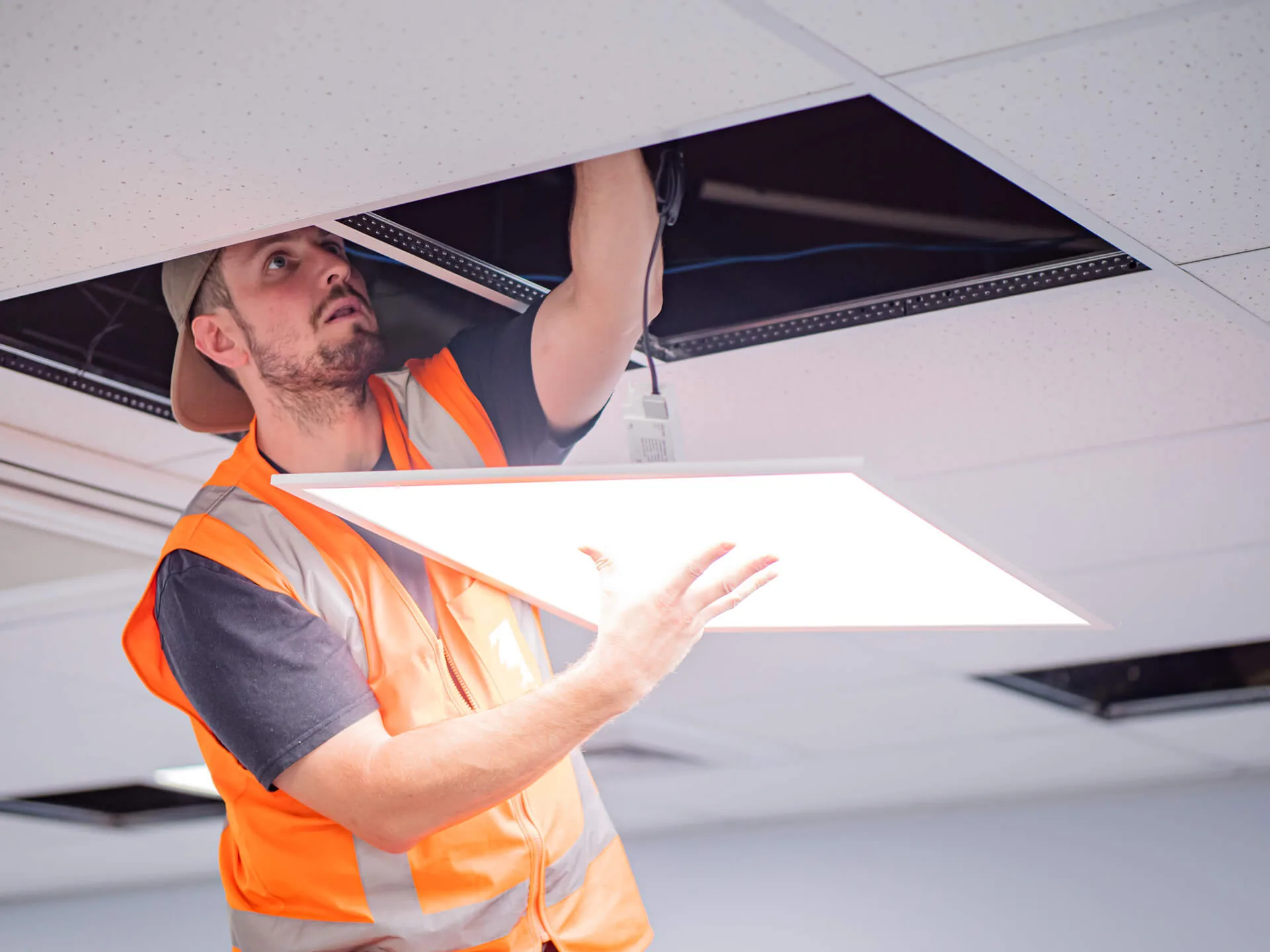 An electrician in a hi vis vest fitting an LED panel into a suspended ceiling